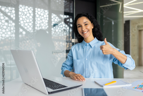 Successful business woman at workplace inside office, hispanic woman smiling and looking at camera, showing thumbs up affirmatively, satisfied with achievement financial results.