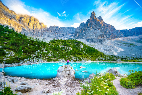 Athletic woman enjoys beautiful view from huge boulder on turquoise Sorapis lake with dito di dio in the evening. Lake Sorapis, Dolomites, Belluno, Italy, Europe.