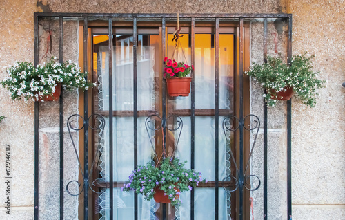 Ventana con cristal y rejas donde cuelgan unas macetas con plantas en flor. Casa de arquitectura moderna, pero tradicional en Hoyos del Espino, Ávila, España. 