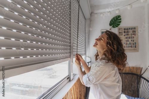 Young woman opening roller blind shutters on the balcony on a summer day