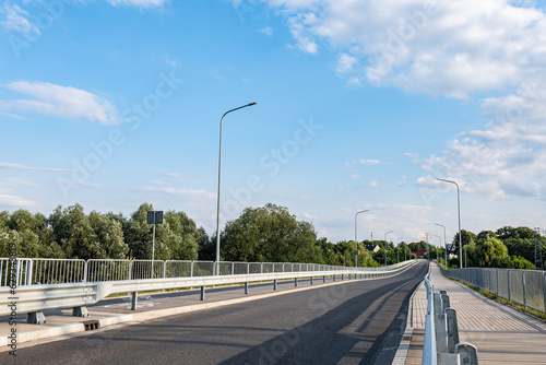 Oświetlenie drogi w obszarze wiejskim, na pierwszym planie barierki ochronne, w tle drzewa i błękitne niemalże bezchmurne niebo