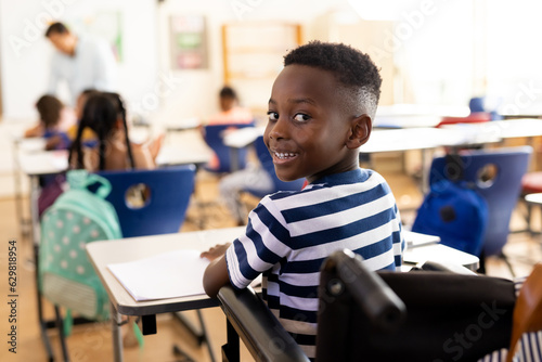 Portrait of happy african american school boy sitting in wheelchair in class at elementary school
