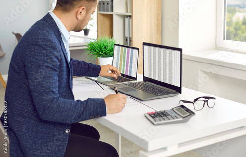 Man in office working with big data and databases using two laptops and excel tables. Office worker making analysis and report with spreadsheets on computer sitting at table near calculator.