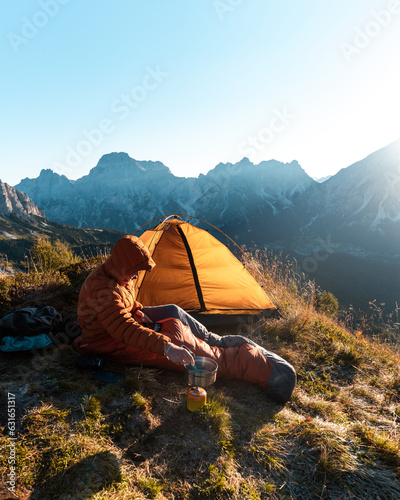 Camping in the wilderness. Man outside his tent is cooking breakfast during the sunrise in the mountains.
