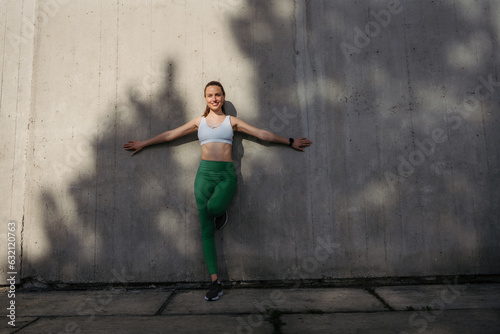 Portrait of young smiling woman in sportswear leaning against the concrete wall in the city.