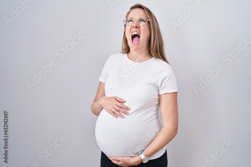 Young pregnant woman expecting a baby, touching pregnant belly angry and mad screaming frustrated and furious, shouting with anger looking up.