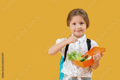 Happy little girl with backpack pointing at lunchbox on yellow background
