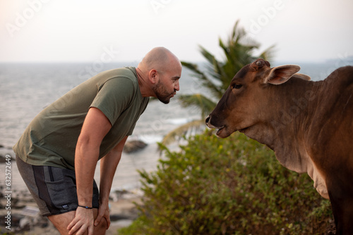 Friendly vacationist leaning towards humped cattle standing against seascape and meeting brown zebu, looking at animal.