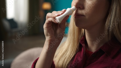 Blonde woman sitting on the sofa, spraying a nasal spray into her nose