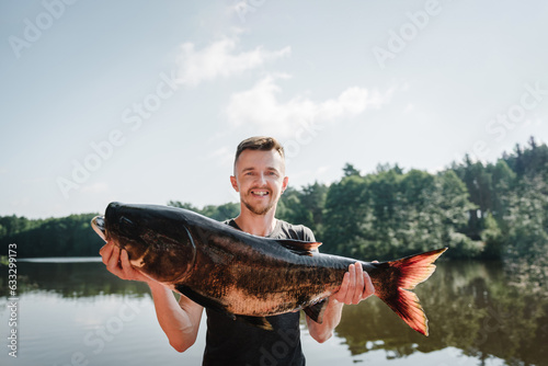 Happy fisherman hold big trophy fish near lake. Success pike fishing. Fresh fish trophy in hands. Young man returning with freshly caught fish. Article about fishing day. Closeup. Fishing backgrounds.