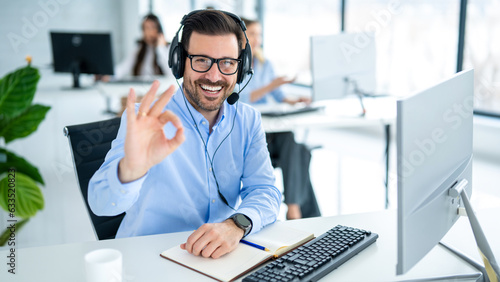 Male customer service representative showing approval sign to the camera, smiling and gesturing okay at call center office.