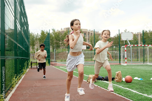 Active schoolgirl in white sportswear leading in running competition while moving along race track with intercultural classmates on background
