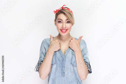 Portrait of blonde woman making thumbs up sign, demonstrates support and respect to someone, smiles pleasantly at camera, achieves desirable goal. Indoor studio shot isolated on gray background.