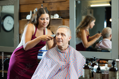 Young woman barber using clippers while providing professional haircut to relaxed elderly man seated comfortably in chair in barbershop ..