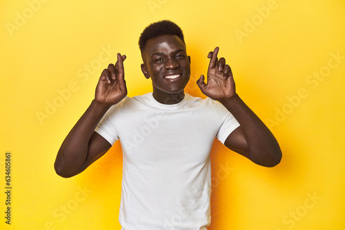 Stylish young African man on vibrant yellow studio background, crossing fingers for having luck
