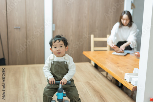 春の昼に部屋で三輪車にまたがって正面を見る日本人の子と母親