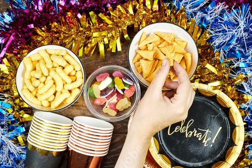 birthday or christmas, new year's party concept, paper plate with card celebrate text and disposable cup, straws and colorful with different chips and candies, pink hat tinsel on wooden table