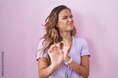 Young hispanic woman standing over pink background disgusted expression, displeased and fearful doing disgust face because aversion reaction.