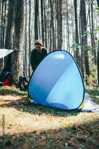 Asian male hiker is setting up a bright blue tent in the forest. Concept of tourism, hiking and staying in nature.