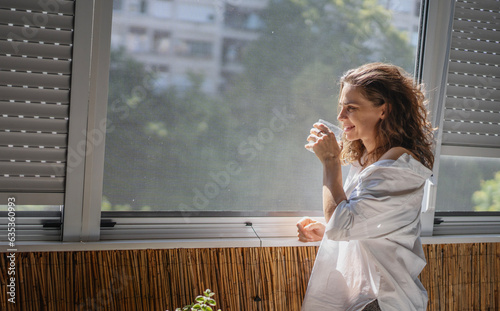 Young smiling caucasian woman in a white shirt standing on the balcony in the morning drinking water and meeting a new day