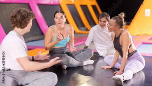Company of four young happy friends enjoying time together while talking and sitting on trampoline in game center indoors