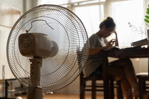 Asian unhappy woman sitting in front of working fan suffering from heat in modern house on sunny summer day.