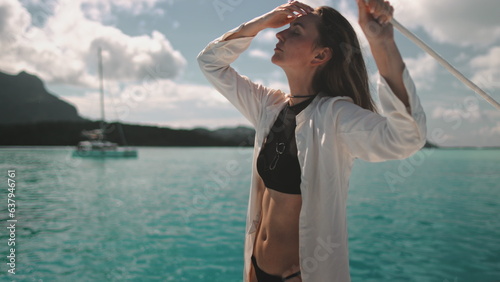 Woman standing on yacht deck looking at the horizon. Turquoise ocean water and mountains in background. Young long hair Caucasian female enjoys travel, tourism, holiday yacht tour to Bora Bora islands