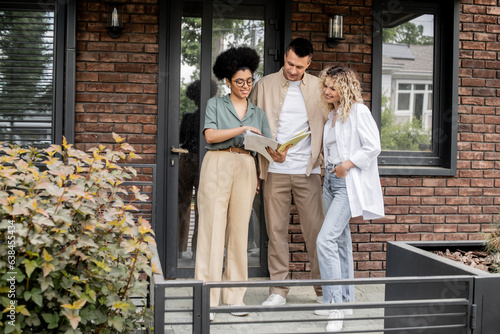 smiling african american real estate agent showing documents to smiling owners of new house in city