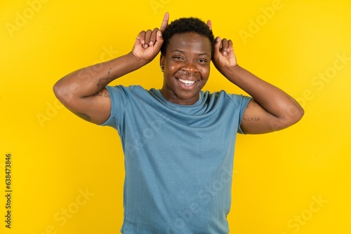 Funny Young handsome man standing over yellow studio background shows horns, fingers on head gesture, posing silly and cute