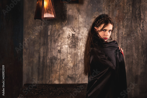 Frowning vampire in cloak posing near chest in underground castle dark room, severe looking at camera. Child girl actress in Dracula image poses indoors. Theatre performing concept. Copy ad text space
