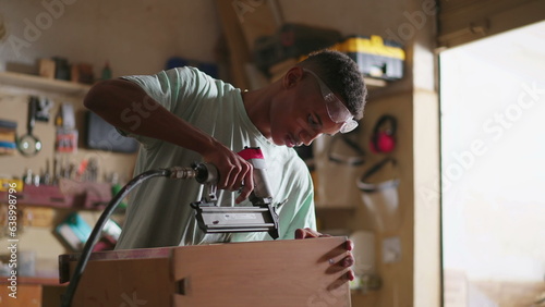 One young black carpenter building and fixing furniture at carpentry workshop. Job occupation of apprentice learning to build wooden object, using drilling equipment