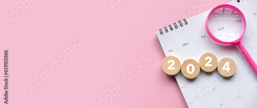 New year 2024 concept. Magnifying glass and calendar with a wooden circle with number over a pink background with copy space.