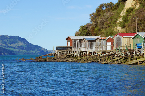 Colourful row of boathouses and boat ramps in a Devauchelle Bay surrounded by bush-covered hills, Onewa, Banks Peninsula, New Zealand.