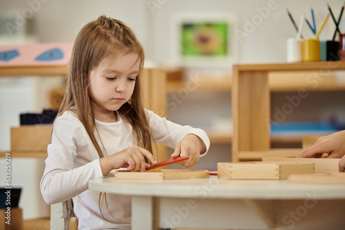 girl playing with wooden didactic materials near friend in class in montessori school