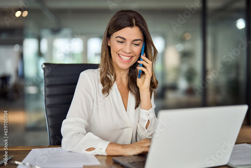 Happy smiling mature middle aged business woman, 40s professional lady executive manager talking on the phone making business call on cellphone at work in office using laptop computer.