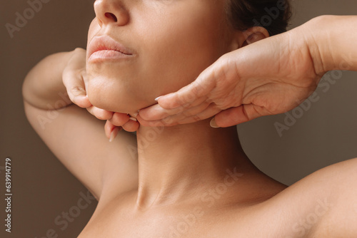 Cropped shot of young caucasian woman touching under the chin with hands massaging her face on dark brown background. Rejuvenation, facelift, facefitness. Exercises from the second chin, pelican neck