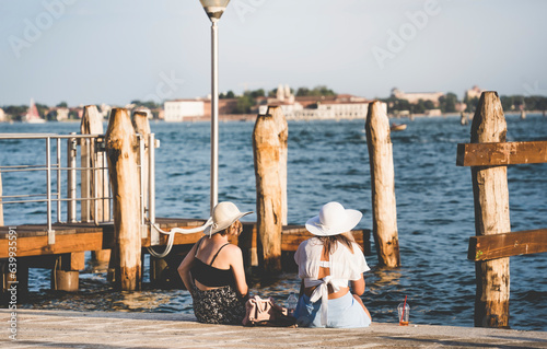 Two tourist girls sitting on the waters edge and enjoying the idyllic scene on Grand Canal in Venice.
