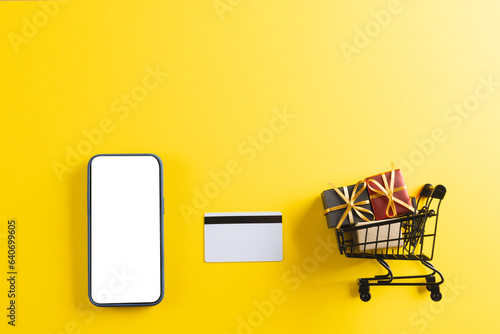 Credit card smartphone with copy space and trolley with gifts on yellow background
