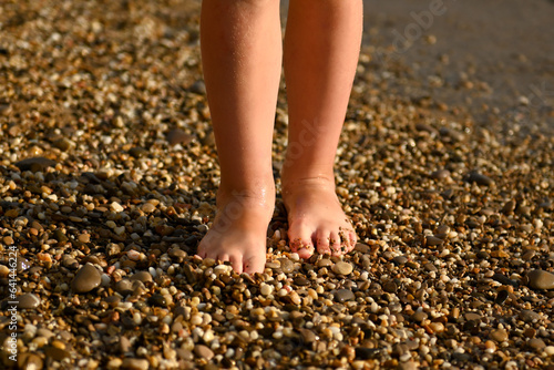 The legs of a boy or girl on the sand of the beach, covered with sand and beach pebbles. Children's feet close-up in small pebbles on the seashore. Children's vacation at the sea. Barefoot beach walks