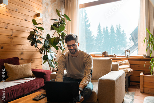 Handsome young adult using laptop while sitting indoors. Playful man surfing the internet in wooden cottage during winter holidays.