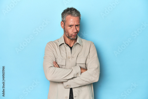 Middle-aged caucasian man on blue backdrop suspicious, uncertain, examining you.