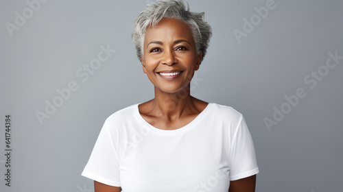 Cheerful mature African American woman with grey hair in casual clothes with a genuine smile on gray background, lookinh at camera in the studio