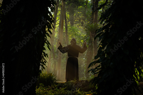 Monje fraile franciscano religioso católico, cristiano meditando o rezando y contemplando la naturaleza en el bosque lleno de árboles y pinos en la neblina y en el amanecer al aire libre