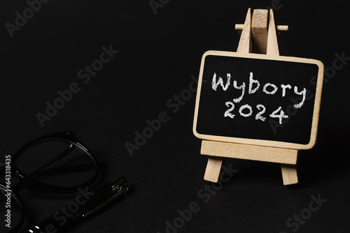 a black writing board on a wooden stand and a black background, the inscription "WYBORY 2024" on it (selective focus)