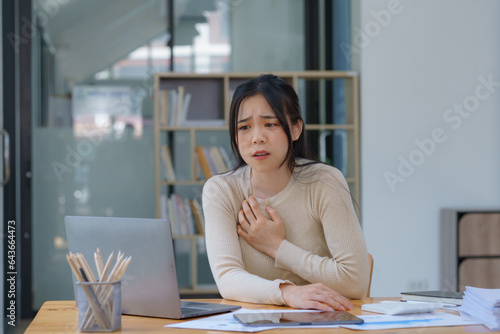 An Asian female has a heart attack pain, and tension-sick employee who is feeling heartache while working on a laptop at the workplace at a desk in the office.