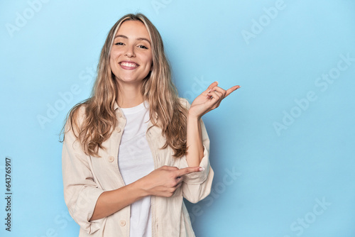 Blonde young caucasian woman in blue studio excited pointing with forefingers away.