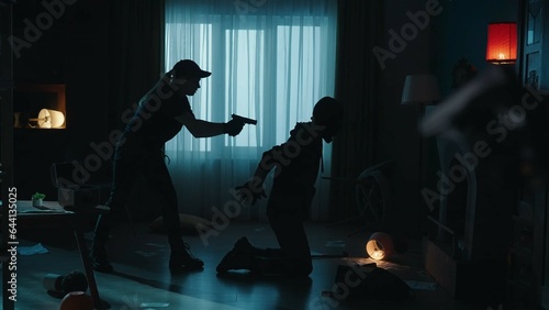 The arrest of a robber in a dark apartmen. Side view of a criminal in a black balaclava, kneeling at gunpoint with his hands behind his back. A female police officer holds the villain at gunpoint.