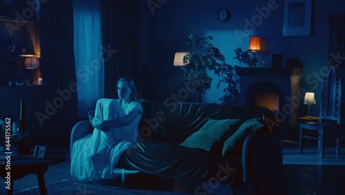 Shot of a young woman sitting on the couch, embracing, hugging her legs and mumbling, whispering something as if she is posessed by a ghost, demon, poltergeist.