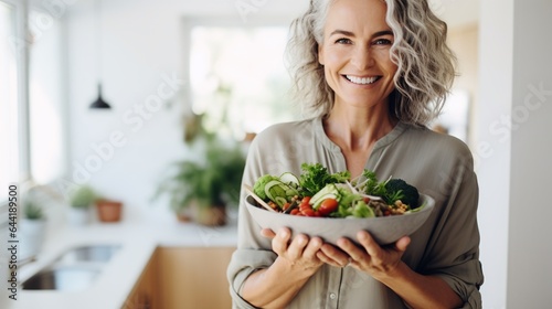 Aged woman smiling happily and holding a healthy vegetable salad bowl on blurred kitchen background, with copy space.