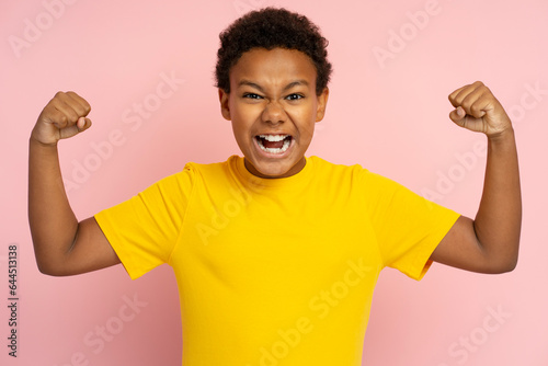 Strong excited African teenager wearing t shirt showing muscles, biceps isolated on pink background
